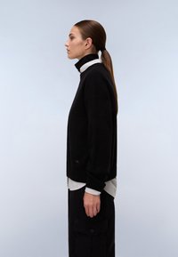 Maglione a maglia nera con colletto alto, indossato sopra una camicia a righe bianche e nere, abbinato a pantaloni cargo neri. Tessuto morbido e vestibilità rilassata.