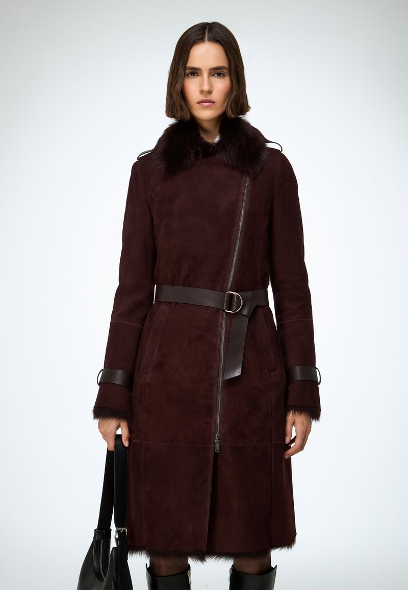 Kabát z bordovej semiše so širokým kožušinovým golierom, zapínaním na zips, opaskom v páse a zdobenými manžetami. Hladká textúra s čistým siluetou.