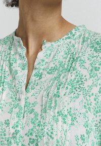 White blouse with green floral pattern, featuring a relaxed collar and button details, textured fabric with subtle pleats.
