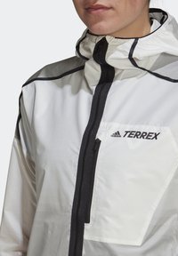 Gros plan d'une personne portant une veste blanche Adidas Terrex avec des détails de fermeture éclair noirs et une poche poitrine avec logo.