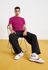 T-shirt fucsia a girocollo, pantaloni neri larghi e sneakers bianche con dettagli bordeaux. Seduto su una sedia in cromo contro un pavimento giallo.