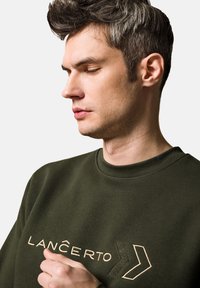 Zielona bluza z okrągłym dekoltem, z haftowaną nazwą marki "LANCERTO" oraz geometrycznymi akcentami w odcieniu beżowym. Gładka tekstura i nowoczesny design.