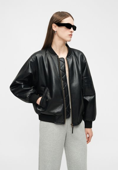 Femme portant une veste bomber en cuir noire oversize, un pantalon gris, des lunettes de soleil rectangulaires noires et des boucles d'oreilles argentées en forme de créoles, tournée vers la droite.