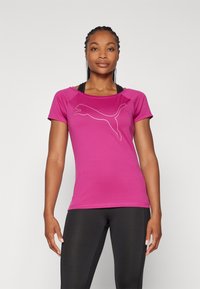 Egy nő rózsaszín, testhez simuló Puma pólót és fekete leggingset visel, ellazított karokkal áll egy sima fehér háttér előtt.