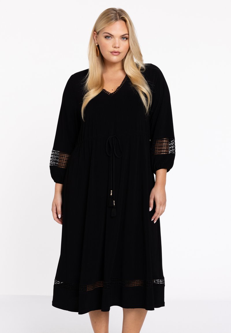 Schwarzes Maxikleid mit V-Ausschnitt und Kordelzug in der Taille. Mit durchscheinenden Spitzeakzenten an den Ärmeln und dem Saum. Hergestellt aus einem weichen, fließenden Stoff.