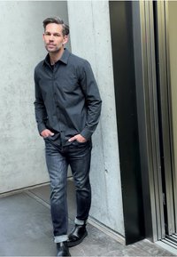 Uomo con camicia grigio scuro a bottoni con un motivo sottile, abbinata a jeans scuri e stivali di pelle neri, che cammina vicino a un ascensore.