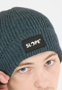 Stickad mössa i mörkblått med ribbad textur, som har en rektangulär svart etikett med "SLOPE"-logotypen i vitt.