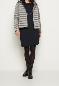 Veste matelassée grise avec capuche, robe bleu marine, collants noirs et bottines. La veste a une texture matelassée et une fermeture éclair à l'avant.