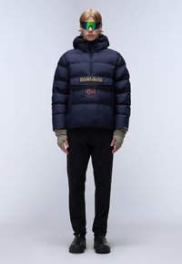 Giacca puffer blu navy con cappuccio, tasca frontale, patch con logo e dettaglio della bandiera norvegese. Pantaloni e guanti neri, occhiali da sole specchiati completano il look.