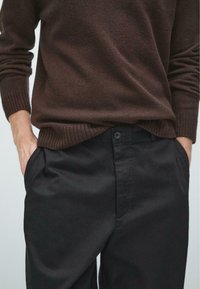 Brązowy sweter z prążkowanymi mankietami, połączony z czarnymi spodniami z zapięciem na guziki i bocznymi kieszeniami, wykonanymi z gładkiego materiału.
