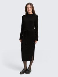 Robe en maille noire avec des manches longues, une ceinture à la taille, des fentes sur les côtés et une longueur midi. Portée avec des mocassins noirs et des collants transparents.