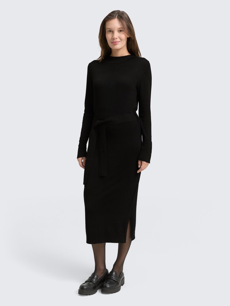 Robe en maille noire avec des manches longues, une ceinture à la taille, des fentes sur les côtés et une longueur midi. Portée avec des mocassins noirs et des collants transparents.