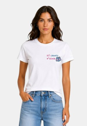 Donna che indossa una maglietta bianca con scritto "it's shorts o'clock" e un piccolo motivo grafico di pantaloncini, abbinata a jeans azzurri chiari.