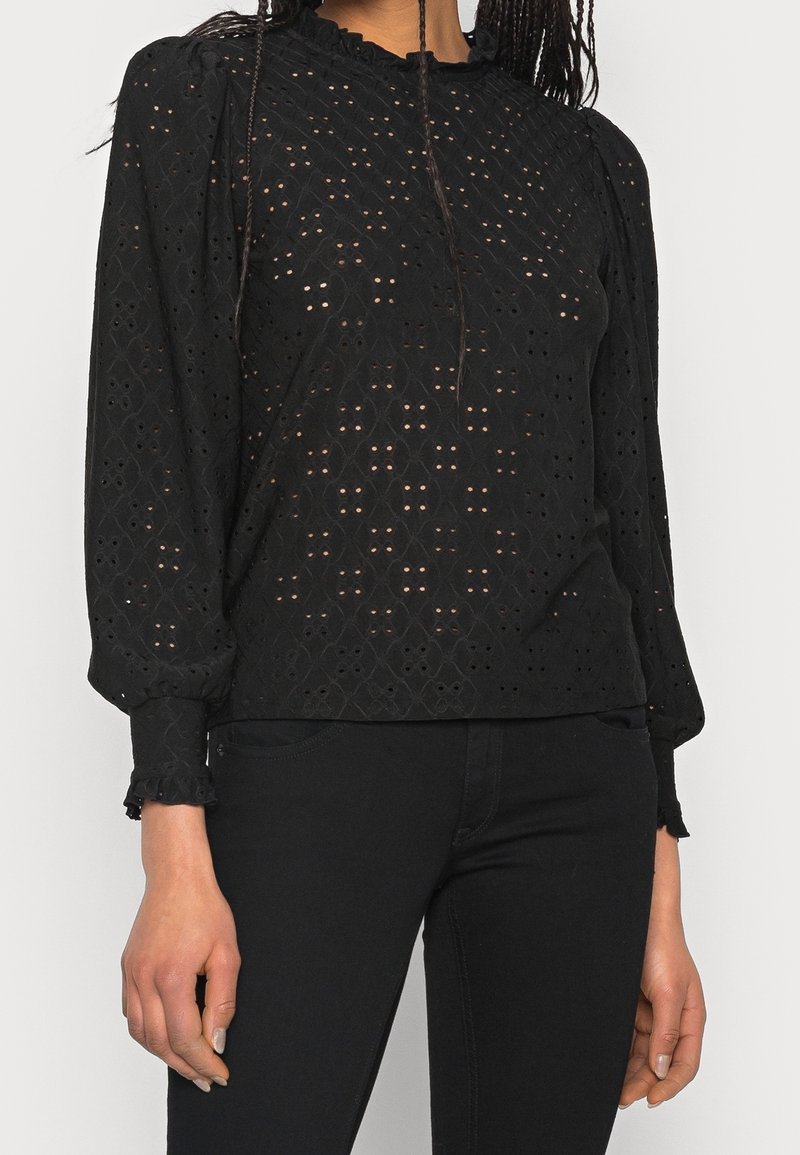 Blouse noire à manches longues en tissu ajouré à motif, avec un col froncé et des poignets volantés. Portée avec un jean noir.