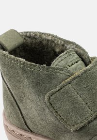 Stivaletto alla caviglia in suede verde con una morbida fodera interna. Presenta una fascia in Velcro e dettagli di cucitura sulla parte superiore e sulla suola. Punta arrotondata.