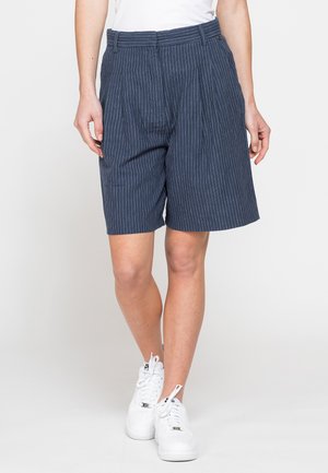 Persona che indossa pantaloncini blu navy a righe sottili fino al ginocchio e scarpe da ginnastica bianche, in piedi davanti a uno sfondo chiaro e uniforme.
