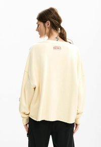 Crème oversized sweatshirt met een onafgewerkte zoom en een rode logopatch op de achterkant. Heeft lange mouwen en een zachte, gestructureerde stof.