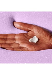 Hand holding a small dollop of white cream on a purple, textured foam background with visible sparkles.