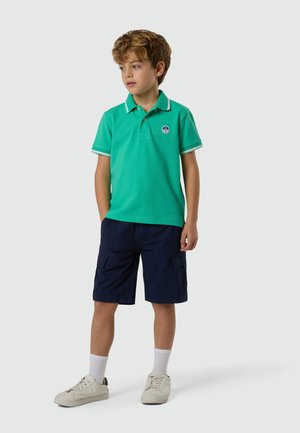 Niño de pie con una camisa polo verde, pantalones cortos cargo azul marino, calcetines blancos y zapatillas blancas, sobre un fondo claro y liso.