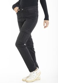 Pantalons cargo en denim noir avec une coupe slim, dotés de poches latérales et d'une fermeture à bouton traditionnelle. Portés avec des baskets blanches.