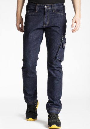 Jeans en denim foncé avec une coupe slim, dotés de coutures jaunes contrastantes et d'une poche latérale. Portés avec des baskets noires et jaunes.