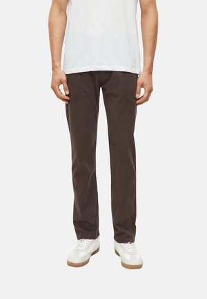 Mann trägt dunkelbraune Hose, weiße Turnschuhe und ein weißes T-Shirt, steht vor einem einfarbigen hellen Hintergrund.