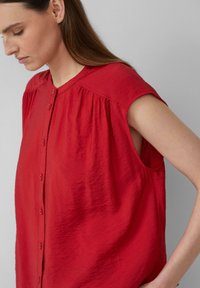 Blouse sans manches rouge avec un devant à boutons, texture douce et froissée, et détail froncé aux épaules.