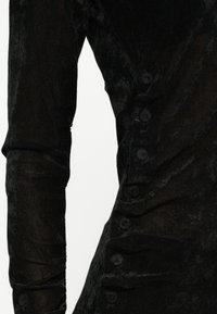 Peça de roupa preta de mangas longas em tecido texturizado, com botões visíveis e detalhes de franzido discretos ao longo do lado. Apresenta seções semi-transparentes.