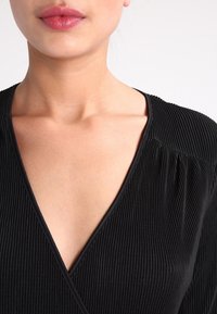 Svart plisserad klänning med djup V-ringning, med texturerad tyg och samlade detaljer vid axlarna.