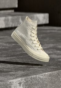 Αθλητικά παπούτσια τύπου high-top από υλικό με υφή σε εκρού απόχρωση, με λείο κάλυμμα δακτύλων, διαθέτουν λευκά κορδόνια και σόλα από καουτσούκ με σχέδιο.