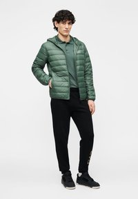 Grüne, gesteppte Steppjacke mit Kapuze, kombiniert mit einem dunkelgrünen Poloshirt und schwarzen Jogginghosen. Schwarze Sneakers mit strukturierter Oberfläche.