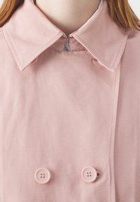 Vaaleanpunainen trenssitakki, jossa kaksirivinen etuosa kahdella napilla, suuri kaulus ja sileä kangaspinta. Vetoketjukiinnitys kaulassa.