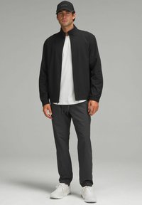 Chaqueta ligera negra con un cuello alto, combinada con una camiseta blanca texturizada y pantalones gris oscuro. Zapatillas blancas completan el conjunto.