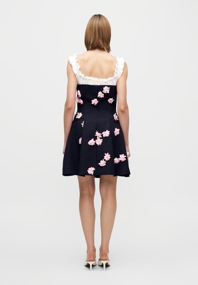 Vestito blu navy con corpetto aderente, spalline bianche arricciate e applicazioni floreali rosa sparse sulla gonna. Linea a trapezio, lunghezza al ginocchio.