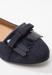 Chaussure plate en suède marine avec un nœud noir brillant et des accents brillants superposés à l'avant. Texture lisse avec une forme arrondie.
