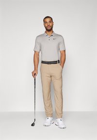 Vīriešu golfa apģērbs, kas sastāv no gaiši pelēka polo krekla, bēšām biksēm, melnas jostas un baltām golfa kurpēm, tur rokā golfu nūju. Vienkāršs dizains, funkcionāls audums.