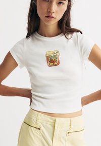 Weißes, kurzärmeliges, cropped T-Shirt mit einer Illustration eines Pfirsichmarmeladenglases, das einen gelben karierten Deckel und ein Bändchen auf der Vorderseite zeigt.