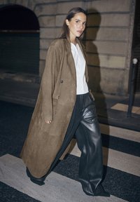 Manteau en daim marron, chemise blanche et pantalon noir à jambes larges texturé, marchant sur un passage piéton la nuit avec un bâtiment en pierre en arrière-plan.