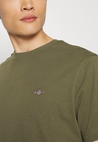 Tricou din bumbac de culoare verde măslinie, cu guler rotund și mâneci scurte, prezentând un mic logo brodat pe piept.