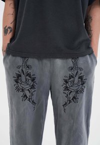 Grijze broeken met zwarte geborduurde ontwerpen van rozen verweven met dolken aan beide zijden, met een gladde stoftextuur.