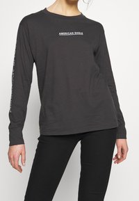 Tmavě šedé tričko s dlouhým rukávem vyrobené z bavlny, s bílým logem "AMERICAN EAGLE" na hrudi a textem podél rukávu. Hladká textura.