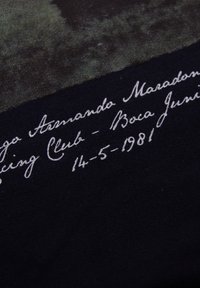 Zwarte stof met een gestructureerd oppervlak. Bevat witte handgeschreven tekst met daarin "Diego Armando Maradona," "Boca Juniors," en de datum "14-5-1981."