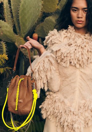 Femme en tenue beige texturée tenant un sac seau marron tressé avec des cordons jaunes, debout devant de grandes plantes de cactus verts.