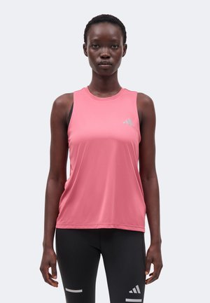 Femme portant un débardeur de sport rose sans manches et un legging noir avec des logos Adidas blancs, debout devant un fond uni.