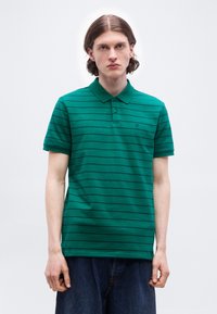 Jeune homme aux cheveux mi-longs portant un polo vert à manches courtes avec de fines rayures noires horizontales et un jean foncé.