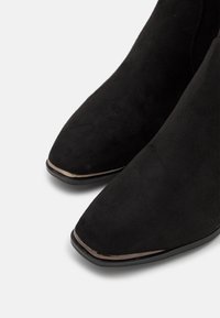 Botines de ante negro con punta afilada, con un ribete negro brillante a lo largo del borde y una silueta elegante.