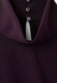 Prenda de color púrpura oscuro con un collar drapeado y tres botones grises en una tapeta negra en la parte posterior del escote.