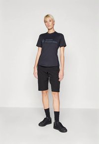 Μαύρο κοντομάνικο πουκάμισο με το λογότυπο "Sweet Protection", συνδυασμένο με μαύρο σορτς. Το μοντέλο φορά μαύρα αθλητικά παπούτσια και κάλτσες, στέκεται μπροστά σε ουδέτερο φόντο.