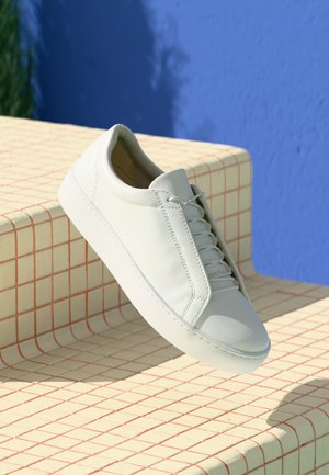 Sneaker bianco in pelle con punta arrotondata, pannello laterale testurizzato e lacci bianchi, posizionata su una superficie a griglia e sfondo blu.