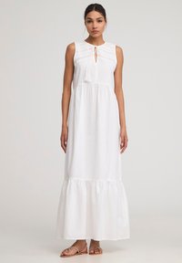 Abito maxi bianco con tessuto strutturato, design senza maniche, scollatura a chiave e orlo a strati. Accentuato da un nodo e frange.